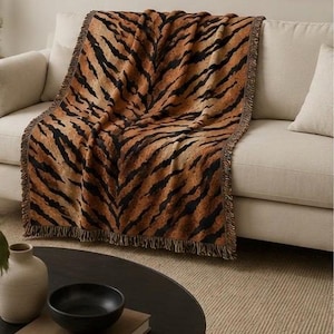 May include: A rectangular throw blanket featuring a prominent tiger stripe pattern in shades of orange, brown, and black. The blanket has a textured surface and is finished with a decorative fringe along the edges, draped over a neutral sofa.