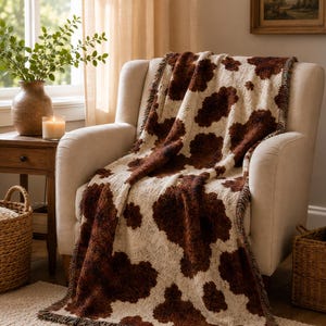 May include: A woven throw blanket featuring a brown and cream cow print pattern with fringed edges. The textile displays a textured surface with irregular dark brown patches distributed across a light cream-colored background fabric.