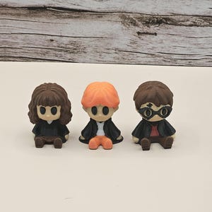 May include: Three small, stylized figurines sitting in a row. The left figure has brown hair and a black robe. The center figure has orange hair, a white shirt, and a black robe. The right figure has brown hair, black glasses, and a black robe.
