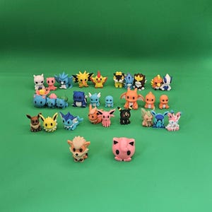May include: A collection of small, colorful plastic character figurines arranged in rows. The set includes various creatures in shades of blue, pink, orange, yellow, brown, and black, featuring distinct shapes, ears, tails, and facial expressions.