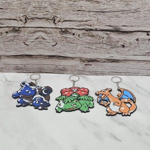 May include: Three pixel art keychains made from fused plastic beads. The left keychain features a blue and brown turtle-like creature. The center keychain depicts a green creature with a large red flower. The right keychain shows an orange dragon.
