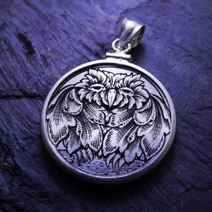 May include: A silver pendant with an intricate engraved design of an owl with its wings spread. The owl is facing forward with its eyes closed. The pendant is on a dark blue background.