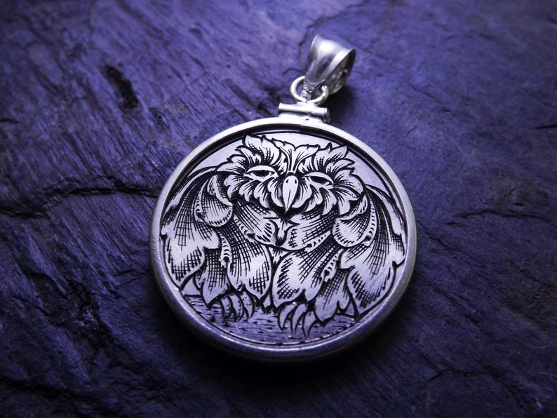 May include: A silver pendant with an intricate engraved design of an owl with its wings spread. The owl is facing forward with its eyes closed. The pendant is on a dark blue background.