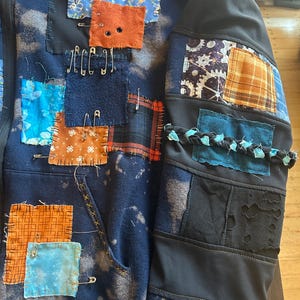 May include: A dark blue bleach-dyed zip-up hoodie featuring numerous fabric patches in various colours and patterns, including orange, blue, and plaid. The patches are attached with visible stitching and silver safety pins, with a braided fabric detail.