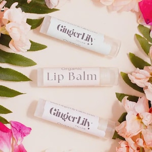 Può includere: Tre tubetti cilindrici di balsamo labbra con etichette bianche. Due tubetti riportano il testo Ginger Lily Tallow e Organic, mentre il tubetto centrale mostra il testo Organic Lip Balm in un carattere serif grigio scuro su sfondo bianco.