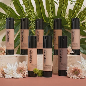 Puede incluir: Ocho frascos cosméticos cilíndricos con bomba etiquetados como Ginger Lily BioGlow en varios tonos de piel. Cada frasco tiene una tapa de bomba negra y un cuerpo transparente que muestra el contenido líquido. Los frascos están sobre bloques de madera clara.
