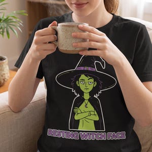 May include: A black short-sleeved t-shirt featuring a graphic of a green-skinned witch with dark hair, a purple-banded hat, and a crescent moon necklace. The witch has crossed arms and the text RESTING WITCH FACE is printed below.