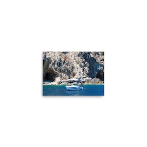 Può includere: Un motoscafo bianco con tettuccio blu che galleggia su acque blu profondo davanti a una grande scogliera rocciosa marrone chiaro e strutturata, con aperture di grotte visibili lungo la costa sotto un cielo luminoso, limpido e soleggiato.