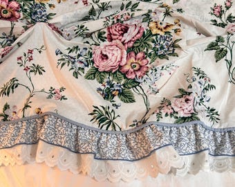 May include: Cream-colored fabric with a floral pattern featuring pink roses, blue and yellow flowers, and green leaves. The fabric has a ruffled edge with a blue and white floral design and a scalloped trim.