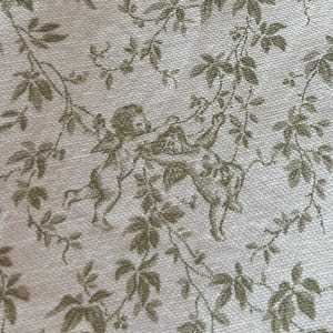 May include: Close-up of a cream-colored fabric with a repeating pattern of olive green leaves and cherub figures. The fabric has a textured appearance, suggesting a linen or similar material.
