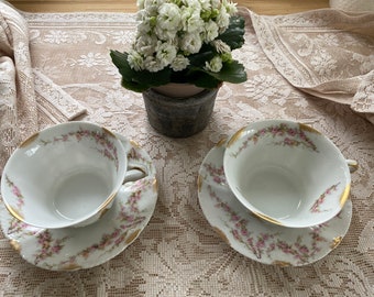 May include: Two white porcelain teacups and matching saucers featuring a delicate pink floral pattern and gold-painted scalloped rims. The cups have curved handles and are arranged symmetrically, showcasing a fine, translucent ceramic finish.