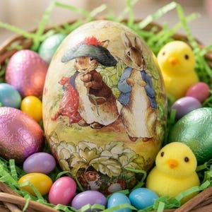 May include: A woven basket filled with Easter decorations. The basket contains a large decorated egg with rabbit illustrations, colorful foil-wrapped eggs, yellow chick figurines, and pastel-colored candies, all nestled in green paper grass.