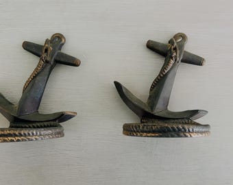 May include: Two bronze-colored anchor-shaped decorative objects. Each anchor has a dark, textured finish with a rope detail and a bronze-colored accent. The anchors are displayed on a light gray surface.