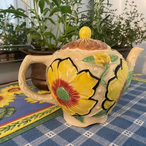 May include: A ceramic teapot with a floral design featuring yellow, red, green, and brown sunflowers. The teapot has a lid and a spout.