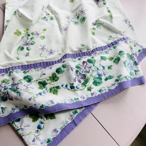 May include: White bed sheet with a floral pattern featuring purple and green flowers and leaves. The sheet has a purple border with a scalloped edge. The flowers are in a watercolor style.