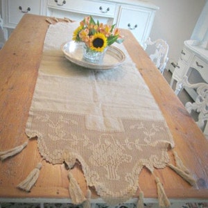 May include: A rectangular beige table runner featuring a decorative crocheted lace border with a floral pattern and hanging tassels at the edges. The runner is centred on a wooden table, providing a neutral base for a silver tray with flowers.