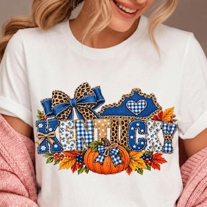 Può includere: Una t-shirt bianca con una grafica della parola KENTUCKY in motivi a quadretti blu e bianchi, stampa leopardata e pois. Il design include un grande fiocco leopardato, una zucca con un fiocco a quadretti blu e foglie autunnali.