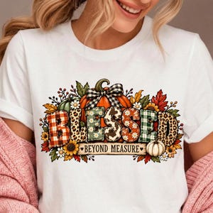 May include: A white t-shirt featuring a graphic of a pumpkin with a black and white checkered bow. The word BLESSED is written in large, patterned letters, with BEYOND MEASURE printed below, surrounded by autumn leaves, flowers, and pumpkins.