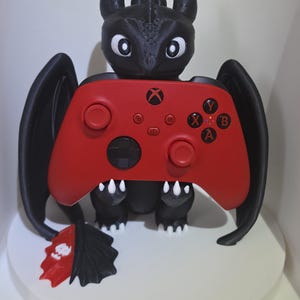 Może przedstawiać: Czarna figurka smoka z dużymi oczami i spiczastymi uszami trzymająca czerwony kontroler do gier wideo. Kontroler posiada logo Xbox, czarne joysticki oraz przyciski oznaczone literami X, Y, A i B. Smok ma białe pazury i czerwoną płetwę ogonową.