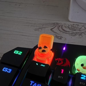 May include: A custom orange mechanical keyboard keycap shaped like a toilet with a lid, featuring a black radiation symbol on the seat. The keycap is illuminated by the keyboard's internal lighting, positioned near the tab key on a black keyboard.