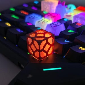 Könnte beinhalten: Eine individuelle mechanische Tastenkappe in Form eines leuchtend roten Würfels mit geometrischem Gittermuster, platziert auf einer schwarzen Tastatur mit hintergrundbeleuchteten Tasten in Blau, Lila, Pink und Gelb.