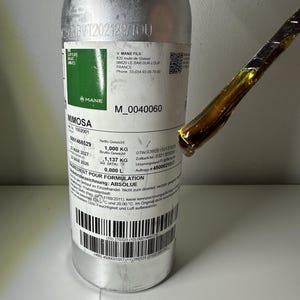 May include: A silver metal bottle labelled Mimosa with a white rectangular label containing technical product information, including a net weight of 1.000 kg and gross weight of 1.137 kg. A metal spatula holds a thick, dark amber viscous liquid.