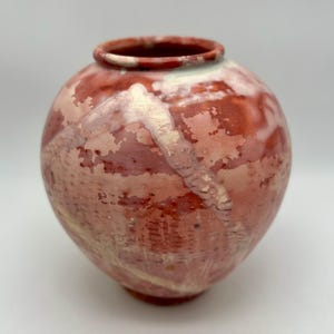 May include: A round ceramic vase featuring a mottled glaze in shades of deep red, terracotta, and creamy white. The surface displays a textured, layered appearance with visible horizontal markings and a smooth, rounded rim opening.