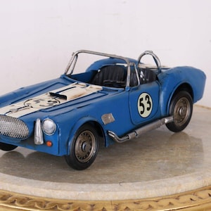 Könnte beinhalten: Ein blaues Metallmodell eines offenen Sportwagens mit einem weißen Rennstreifen auf der Motorhaube, der schwarzen Nummer 53 an der Seite und auf der Haube, einem silbernen Netzgrill, schwarzen Rädern und einem silbernen Überrollbügel hinter den Sitzen.
