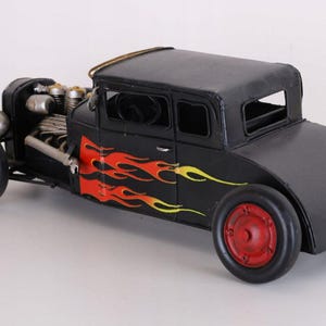 Könnte beinhalten: Ein schwarzes Hot-Rod-Modellauto aus Metall mit einem markanten, freiliegenden Motorblock an der Vorderseite, roten Rädern mit schwarzen Reifen und einer Flammen-Grafik in Rot, Orange und Gelb an der Seite der Karosserie und der Tür.