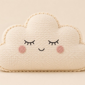 May include: A cream-colored crochet cloud pillow featuring a stitched face with closed eyes and a small smile. The cloud has two circular pink cheeks embroidered on the front and a decorative scalloped edge border around the entire shape.