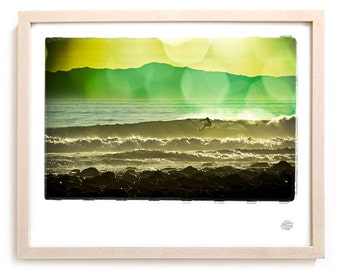 Può includere: Una silhouette di un surfista che cavalca un'onda davanti a un tramonto verde e giallo con una spiaggia rocciosa in primo piano.