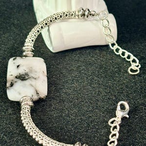 May include: A silver-toned bracelet featuring a rectangular white stone with black inclusions. The bracelet includes intricate metal filigree segments, smooth round spacer beads, and a silver-toned chain with a lobster claw clasp closure.