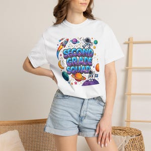 Puede incluir: Una camiseta blanca de manga corta con un gráfico colorido de temática espacial con el texto SECOND GRADE SQUAD. El diseño incluye ilustraciones de astronautas, planetas, un cohete, estrellas y una luna creciente en tonos vibrantes de azul, morado, naranja y rojo.