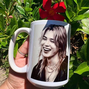 May include: A white ceramic mug featuring a black and white photographic print of a person with long hair, wearing layered necklaces and a dark top, captured in a smiling expression. The mug has a curved handle and a smooth, glossy finish.