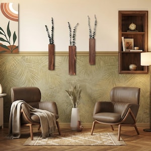 May include: Three rectangular dark brown wooden wall-mounted vases, each holding a sprig of dried eucalyptus. The vases are arranged vertically on a wall above two brown leather armchairs with wooden frames and a white floor vase with dried grass.