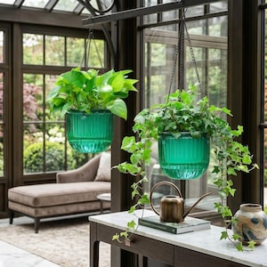 May include: Two hanging planters made of translucent emerald green glass with vertical ribbed textures. Each planter is suspended by a thin metal chain and contains a lush green plant. A small copper watering can sits on a white table.