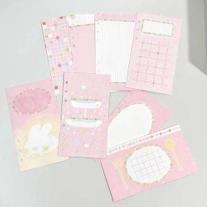 May include: A collection of six rectangular planner inserts in shades of pink. The paper sheets feature various designs including a grid pattern, lined sections, a white rabbit illustration, a tiered fountain, and a dining table setting with utensils.