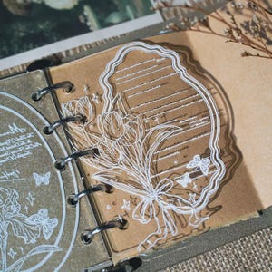 May include: A clear acrylic planner divider featuring a white line art illustration of a tulip bouquet tied with a ribbon, overlaid on a lined note section. The divider has a wavy edge and is secured by silver metal rings in a binder.