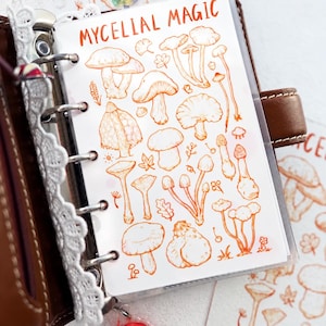 May include: A clear plastic planner insert featuring orange line art illustrations of various mushrooms and the text Mycelial Magic. The insert is held by a brown leather binder with a white lace trim along the metal ring mechanism.
