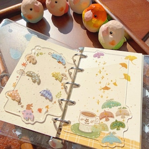 May include: A ring-bound planner with two clear acrylic dividers featuring colorful moth and leaf illustrations, a coffee cup graphic, and ginkgo leaf patterns. Five small, hand-painted wooden bird figurines in pastel colors sit above the planner.