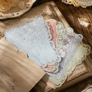 May include: A set of rectangular paper cards with intricate die-cut lace edges in various pastel colours including blue, pink, green, and cream. The cards are partially tucked into a tan paper envelope with the text VINTAGE and have a good day.