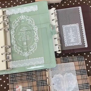 May include: A mint green six-ring binder featuring a clear plastic dashboard with a white lace border and a decorative oval frame containing Spanish text. Beside it, a dark brown binder and a beige plaid binder with a heart-shaped lace dashboard.