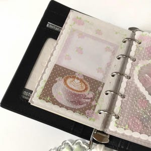 May include: A black six-ring binder featuring a transparent plastic insert decorated with a white polka dot pattern, pink roses, and a central graphic of a coffee cup with latte art on a brown background. The binder has internal card slots.