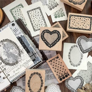 Puede incluir: Una colección de sellos de madera con intrincados patrones de encaje y corazones, acompañada de varios recortes de papel que muestran impresiones en tinta negra de estos diseños, incluyendo motivos ovalados, rectangulares y de corazón.