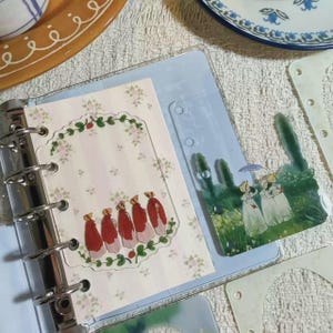 Pode incluir: Uma inserção de fichário de plástico transparente com uma moldura decorativa apresentando cinco figuras em capas vermelhas e capotas, ao lado de um divisor transparente com uma cena pintada de figuras em vestidos brancos com guarda-chuvas.