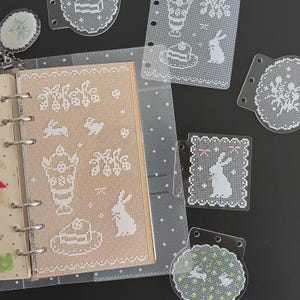 May include: A collection of clear plastic planner inserts and dividers featuring white lace-like patterns of rabbits, cakes, and floral motifs. The items include a rectangular page, several circular and rectangular tabbed dividers, and a small charm.