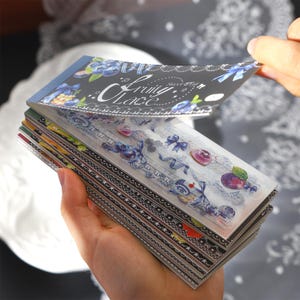 Puede incluir: Una pila de hojas de papel rectangulares con una cubierta negra que presenta el texto Fruity Lace e ilustraciones de arándanos. Las hojas muestran varios diseños que incluyen cintas azules, patrones de encaje y motivos frutales como uvas.