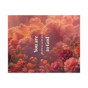 Op de afbeelding: Een rechthoekige legpuzzel met een levendige zonsonderganglucht vol pluizige oranje en roze wolken boven een tuin met bloeiende rode en koraalkleurige pioenrozen. De witte tekst in het midden luidt, You are precious to God.
