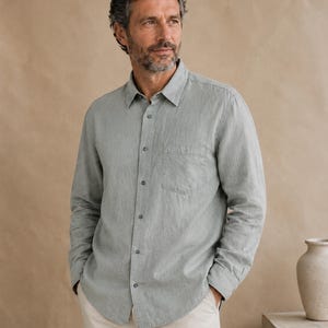 May include: A long-sleeved button-down shirt in a muted sage green color. The shirt features a standard collar, a single chest pocket on the left side, and matching buttons down the front placket. The fabric has a visible, slightly textured weave.