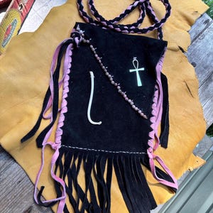 May include: A black suede pouch with pink leather lacing along the sides and a long braided black and pink strap. The front features a white ankh symbol, a curved white bone-like object, and a diagonal strand of small brown beads.